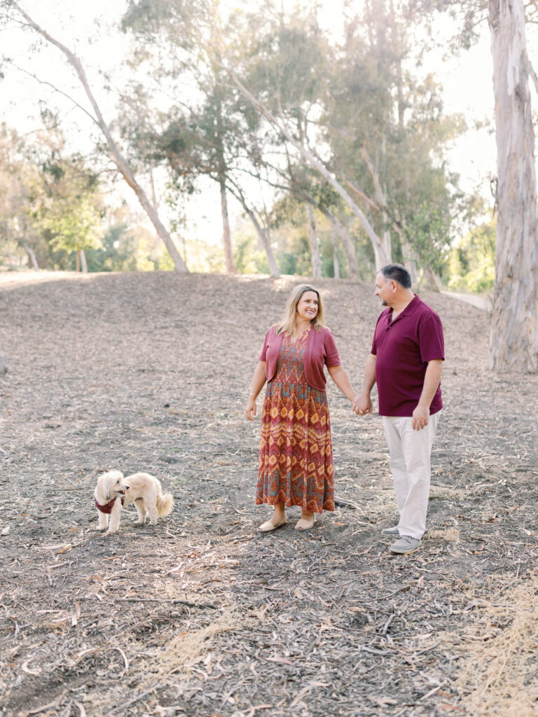 A couple holds hands, smiling at each other in a sunlit forest. They are accompanied by a small dog.