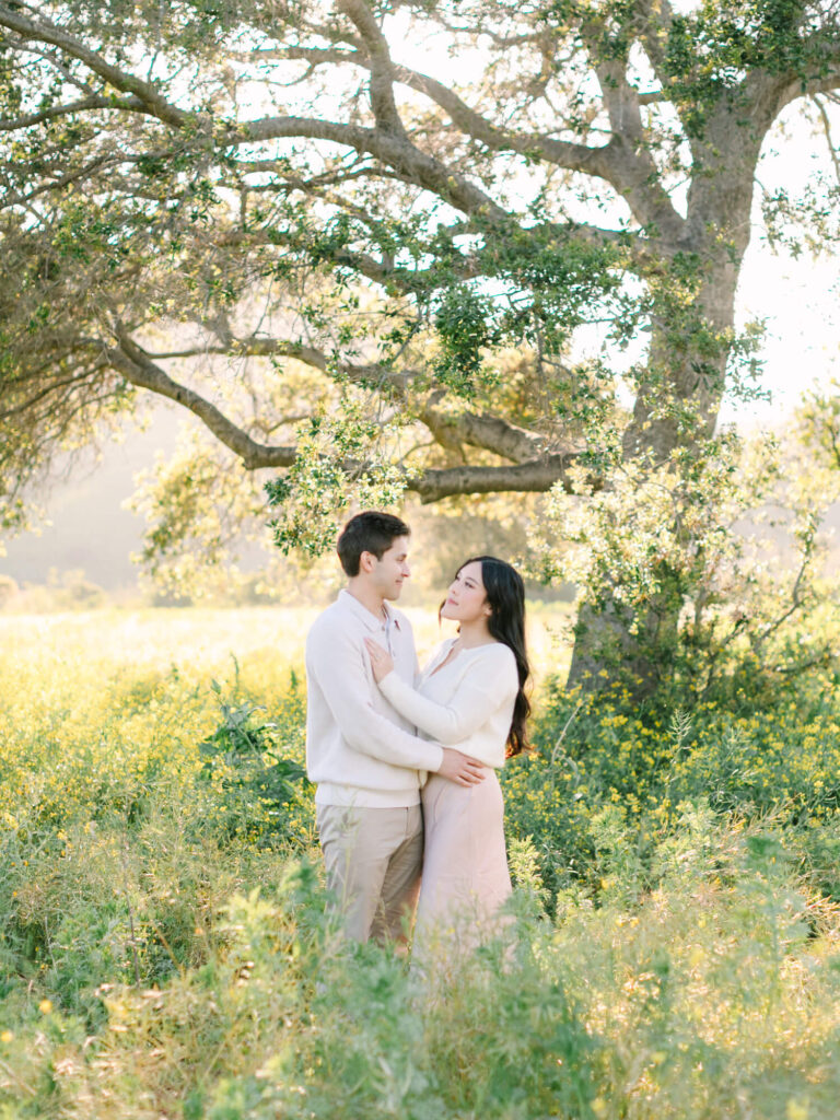 A couple embraces lovingly under a large oak tree in a sunlit meadow. They wear light clothing, surrounded by lush greenery and wildflowers.