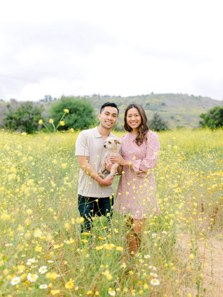 A couple stands smiling in a field of yellow wildflowers, embracing a small dog.