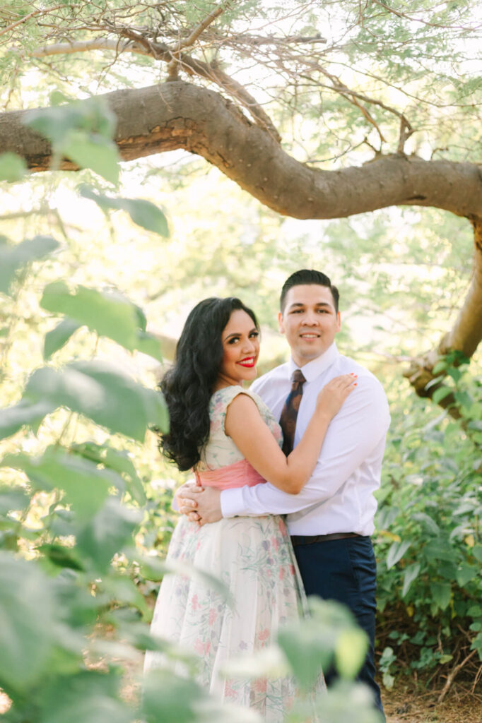 A couple stands smiling under a sunlit tree, surrounded by leafy greenery. The woman in a floral dress embraces the man in a tie.