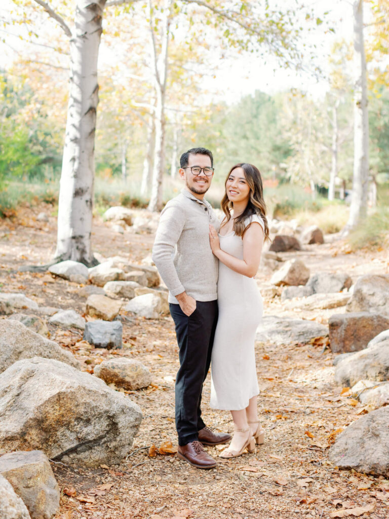 A smiling couple stands in a sunlit forest, surrounded by autumn leaves and rocks.