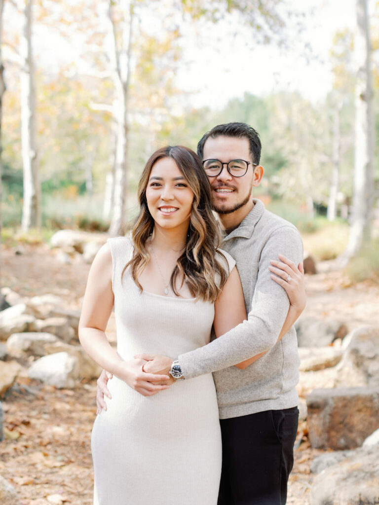A couple embraces in a sunlit park with autumn trees. The woman wears a beige dress; the man, in glasses and a light sweater, smiles warmly.