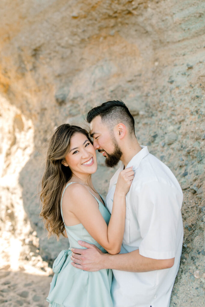 A couple stands close beside a rocky wall, smiling warmly. The woman in a light blue dress looks at the camera, while the man leans in.