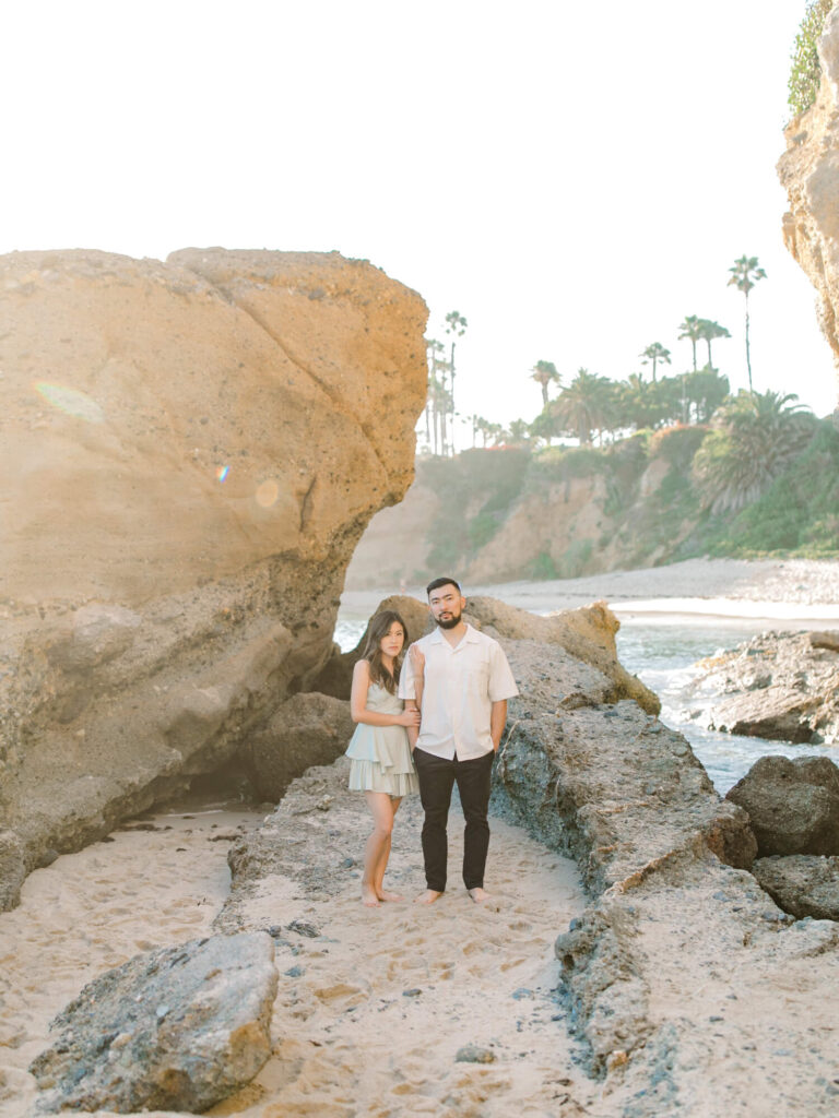 A couple stands on a rocky beach under a clear sky, with sunlight casting a warm glow. Large rocks and cliffs in the background.