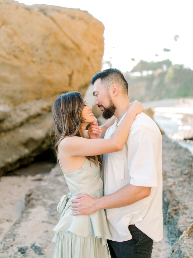 A smiling couple embraces on a sunny beach, standing close to large rocks.