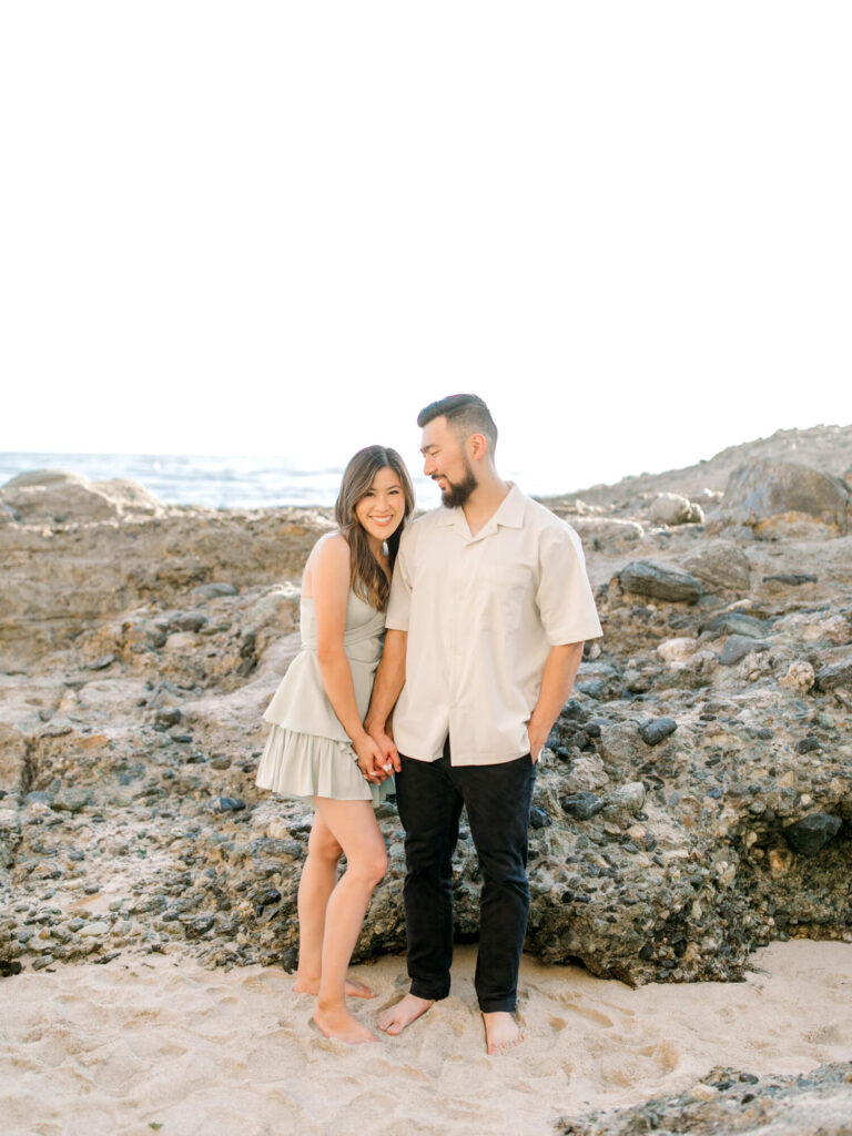 A couple holding hands stands barefoot on a rocky beach, smiling warmly at each other.