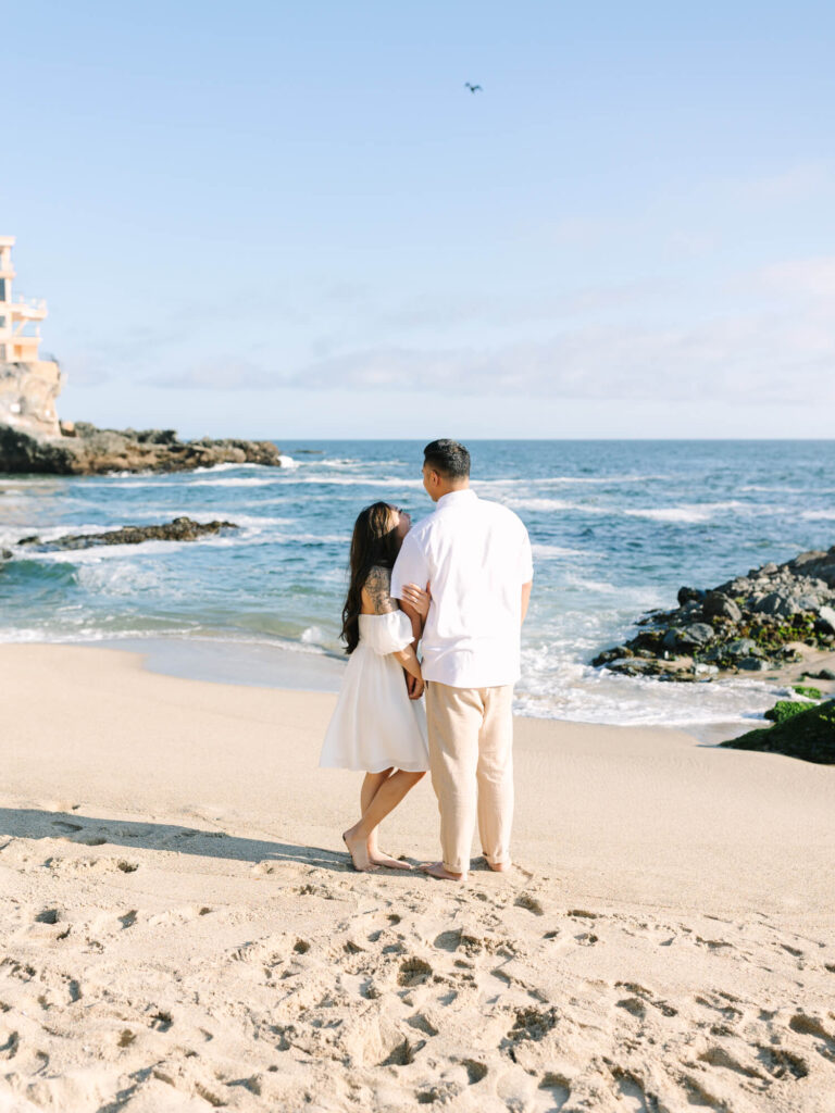 A couple stands closely on a sunny beach, gazing at the ocean.