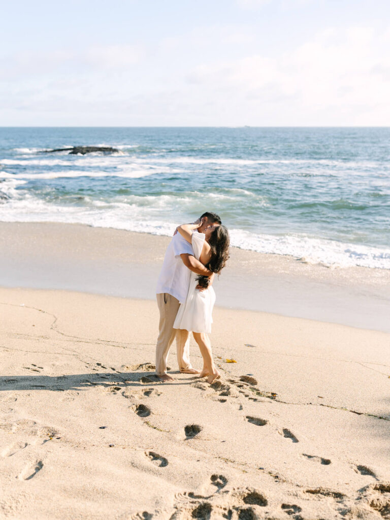 A couple is embracing, with waves gently lapping the shore under a clear blue sky.