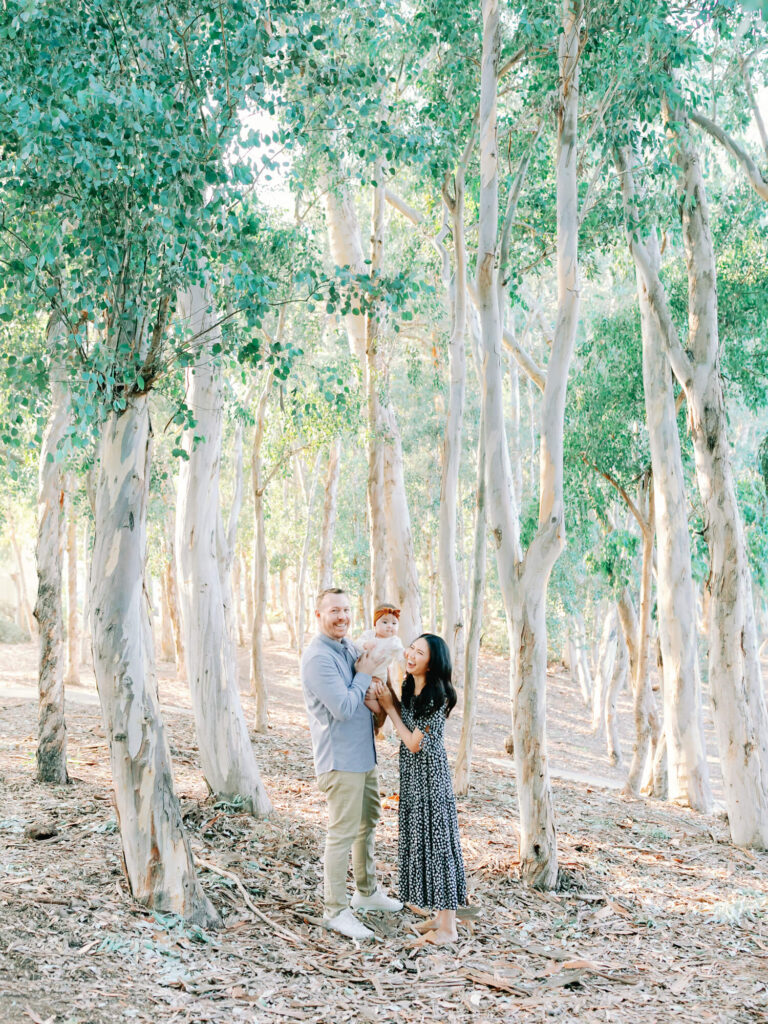 A family of three, standing in a sunlit forest with tall, slender trees.