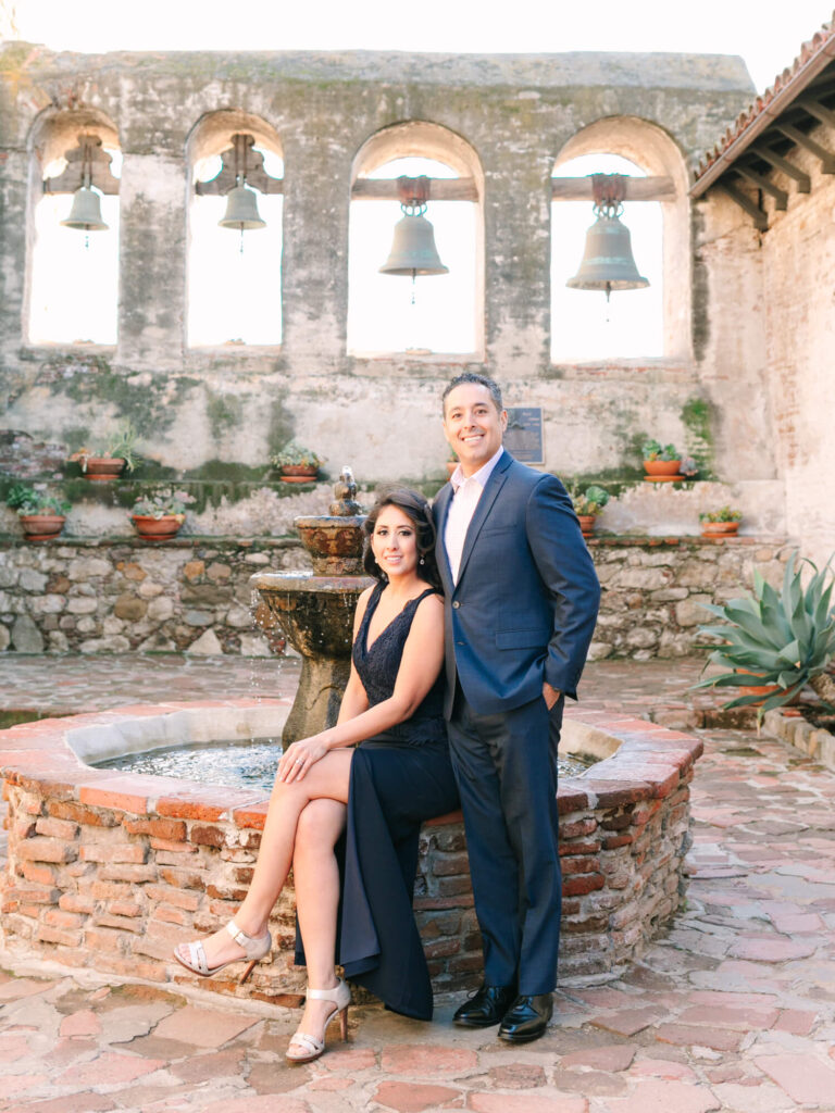 A woman in a navy dress sits gracefully on a brick fountain, beside a man in a blue suit.