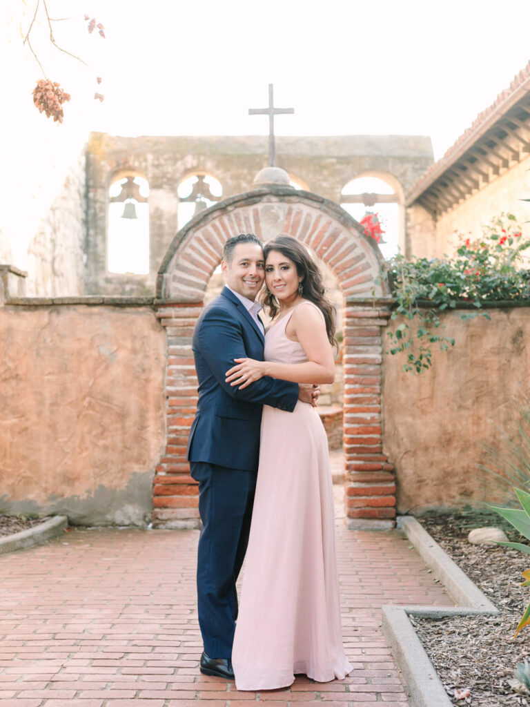 A couple embraces in a sunlit courtyard, surrounded by rustic brick and stone.