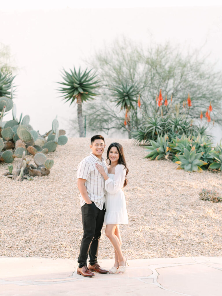 A smiling couple stands on a desert path lined with cacti and agave plants.