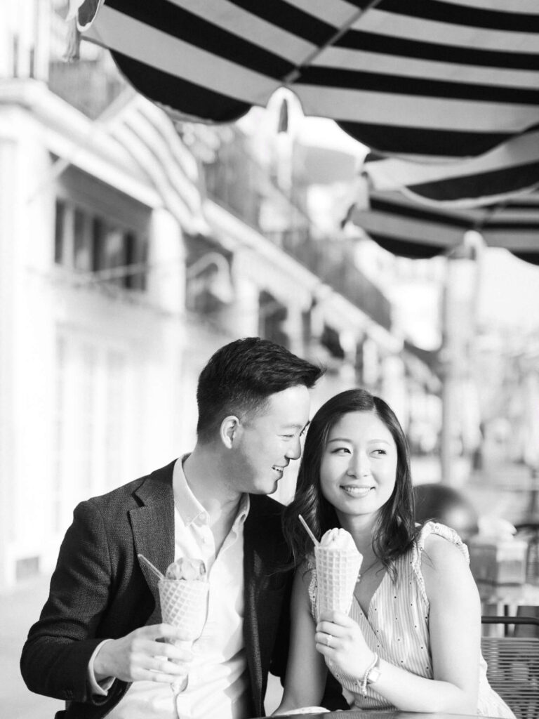 A smiling couple sits under striped awnings, holding ice cream cones on a sunny day.