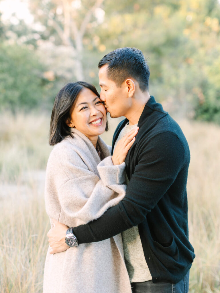 A couple embraces outdoors, with the man gently kissing the woman's forehead. She smiles warmly, wrapped in a beige coat.