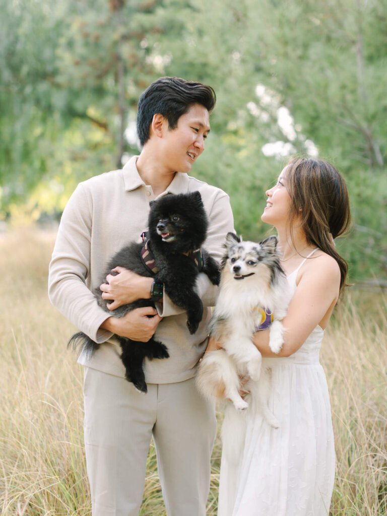 A couple smiles at each other, each holding a small dog. They stand in a grassy field with trees in the background.