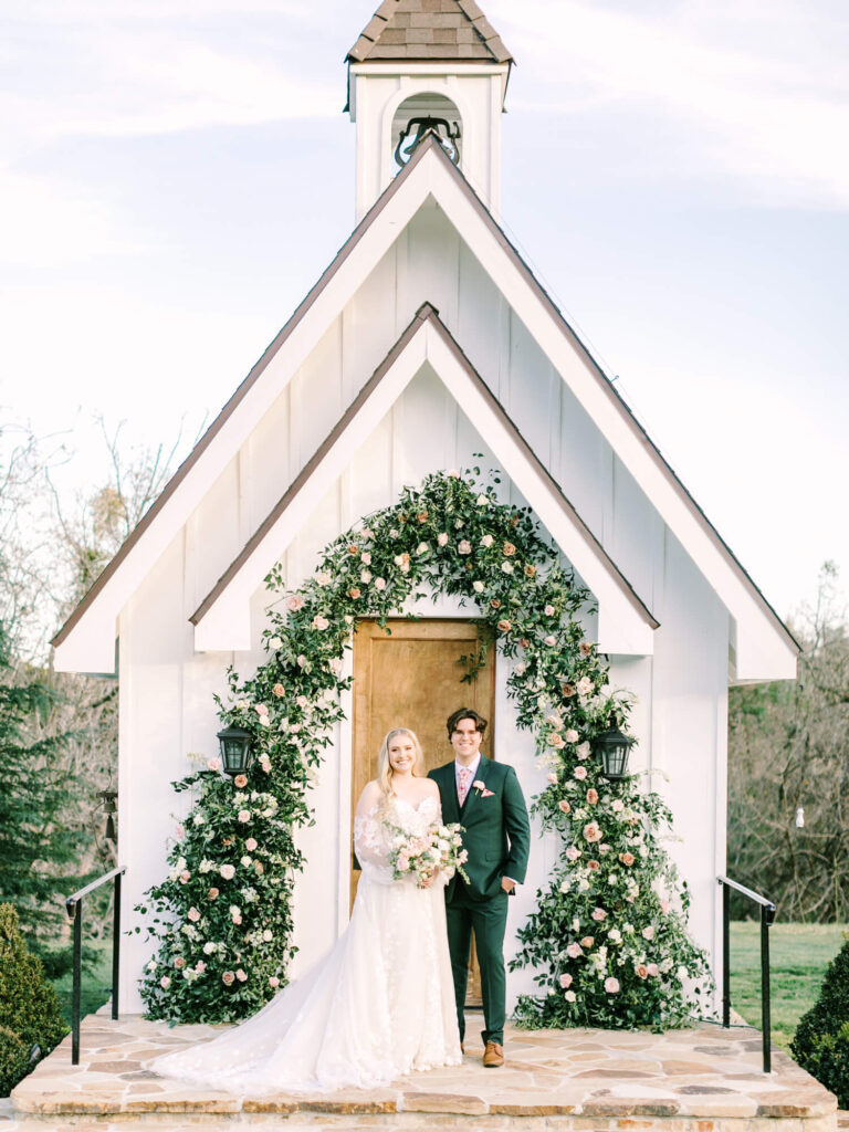 A bride in a lace gown and groom in a dark suit stand happily in front of a small chapel, adorned with a lush floral arch.