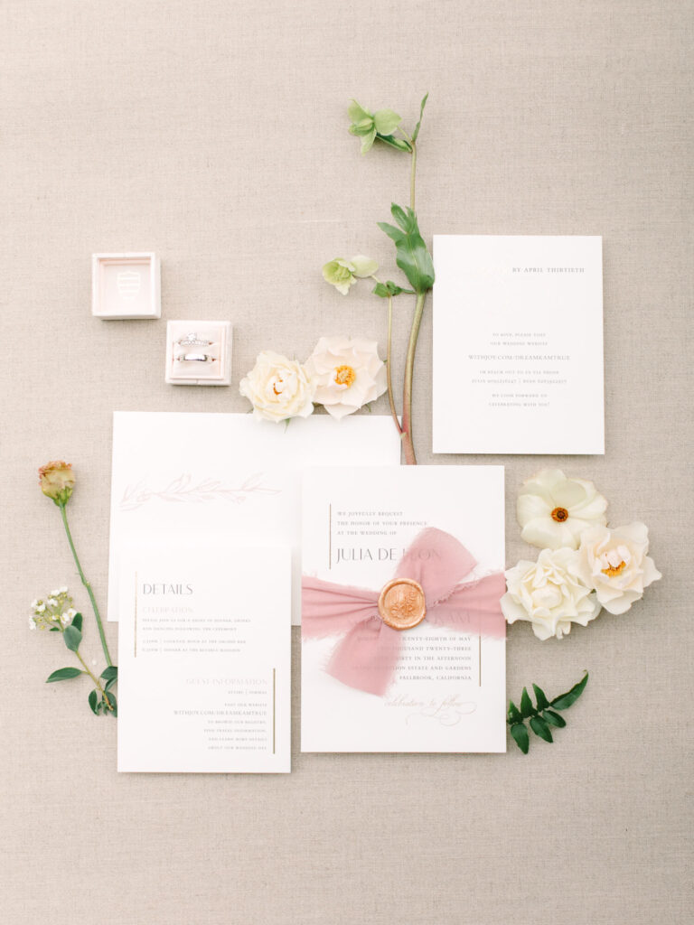 Wedding invitations arranged on a beige fabric background, accented by white flowers and greenery.