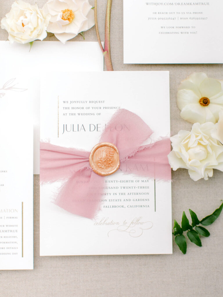 Elegant wedding invitation with a blush ribbon and wax seal, accompanied by white flowers.