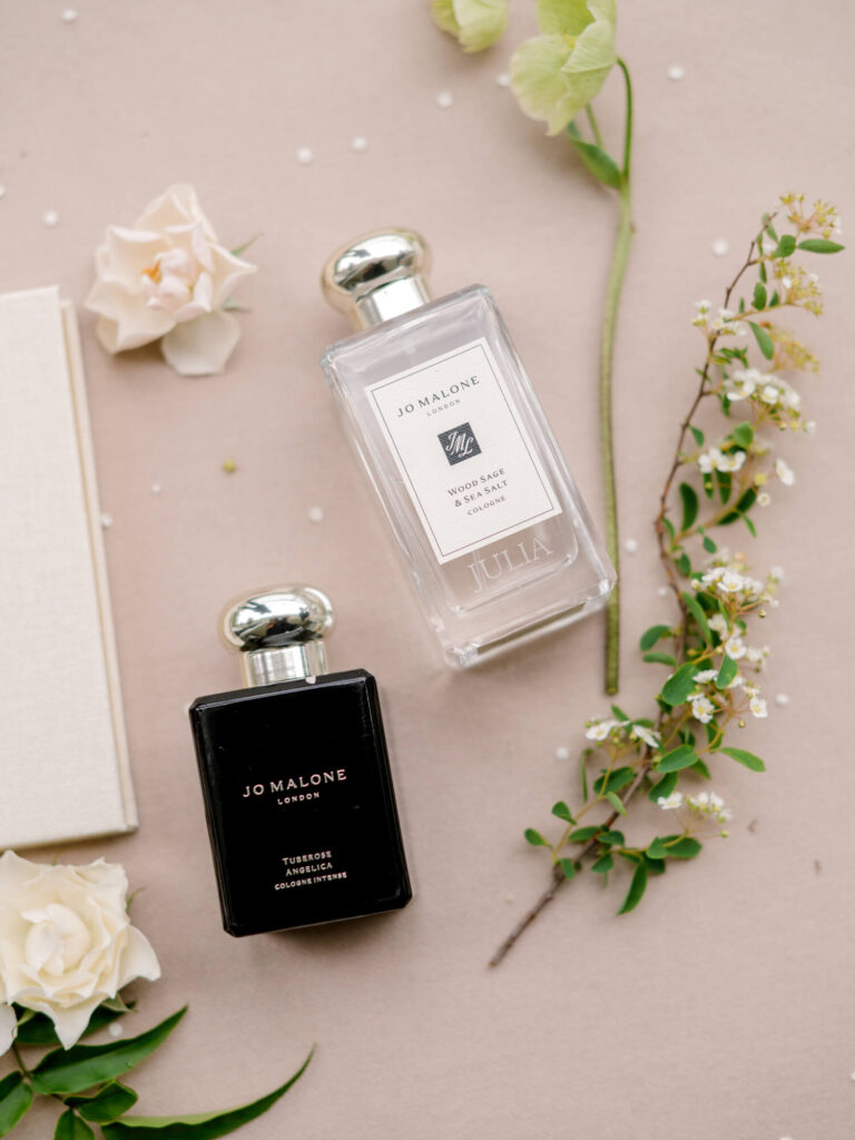Two Jo Malone perfume bottles on a beige background with scattered flowers and greenery. One bottle is black, the other clear.