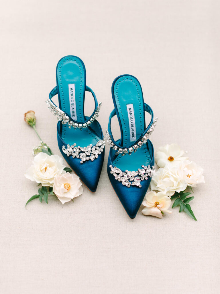 Elegant blue satin heels adorned with sparkling embellishments, surrounded by delicate white flowers on a soft beige background.