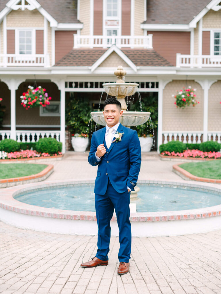 A man in a blue suit stands smiling in front of a fountain, with a charming house and colorful flowers in the background.