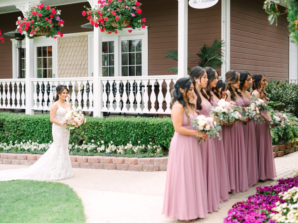 A bride in a lace gown walks toward six bridesmaids in mauve dresses holding bouquets, against a charming porch with hanging flowers.
