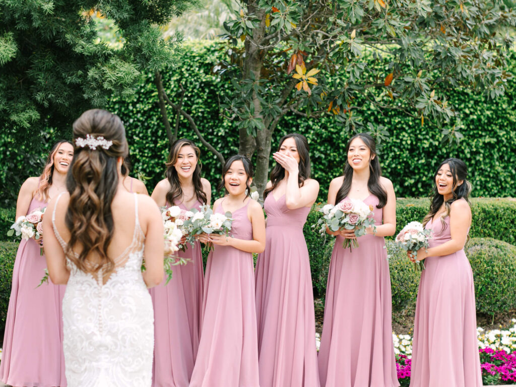 A bride faces six bridesmaids in pink dresses, standing outdoors in front of lush greenery.