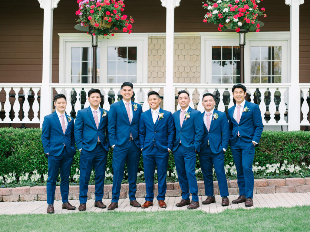 Seven men in matching blue suits with pink ties and boutonnières stand smiling in front of a porch adorned with red and white flower.