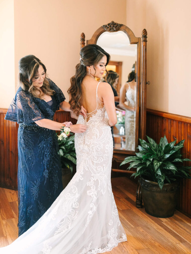 A bride in an elegant white lace gown has her dress adjusted by another woman in a blue dress, both standing in front of a vintage mirror.