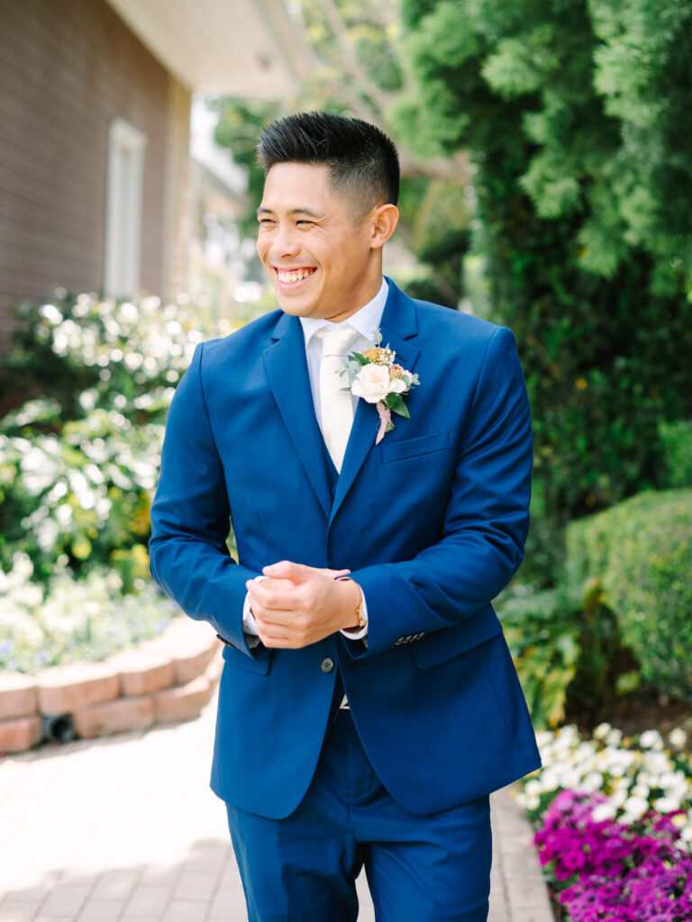 Smiling man in a blue suit with a boutonniere, standing on a sunlit brick path surrounded by lush greenery and colorful flowers, exuding joy.