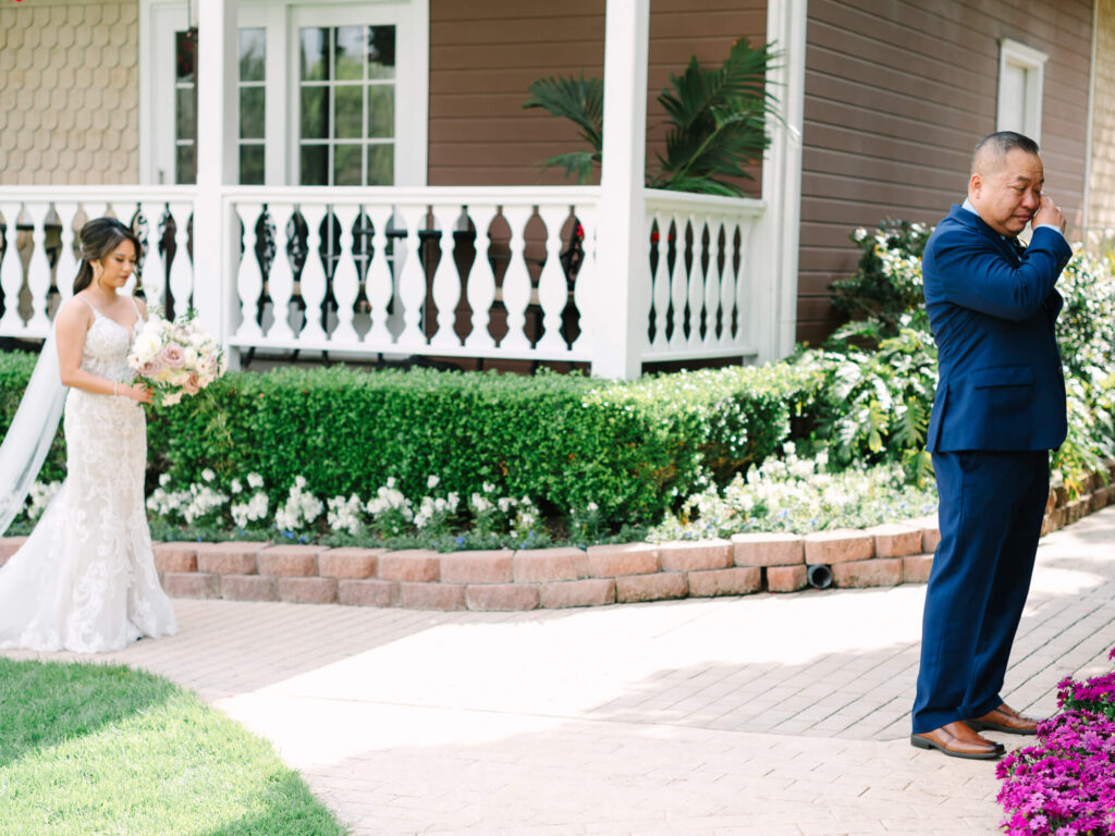 A bride in a white gown approaches her dad in a blue suit, wiping his tear, set against a garden and white porch backdrop.
