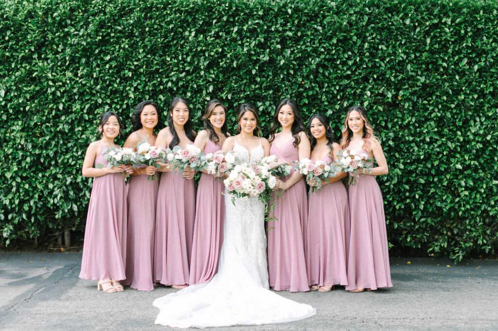 Bride in white gown, surrounded by seven bridesmaids in matching pink dresses, holding bouquets.