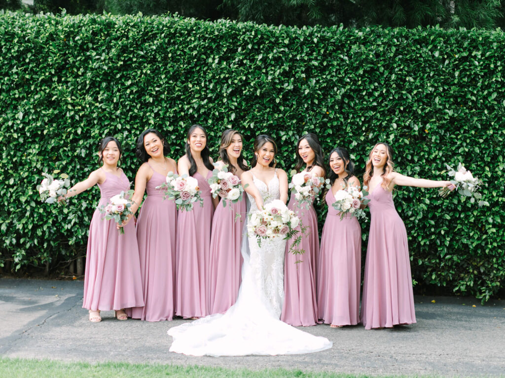 Bride in white gown stands with seven bridesmaids in lavender dresses holding bouquets, all smiling, against a lush green hedge background.