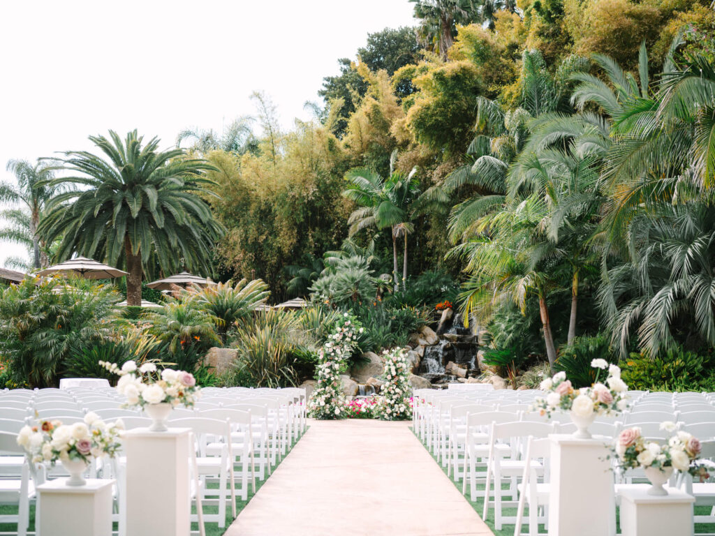 Outdoor wedding setup with white chairs facing a lush tropical garden backdrop. Floral arrangements line the aisle, leading to an archway by a waterfall.