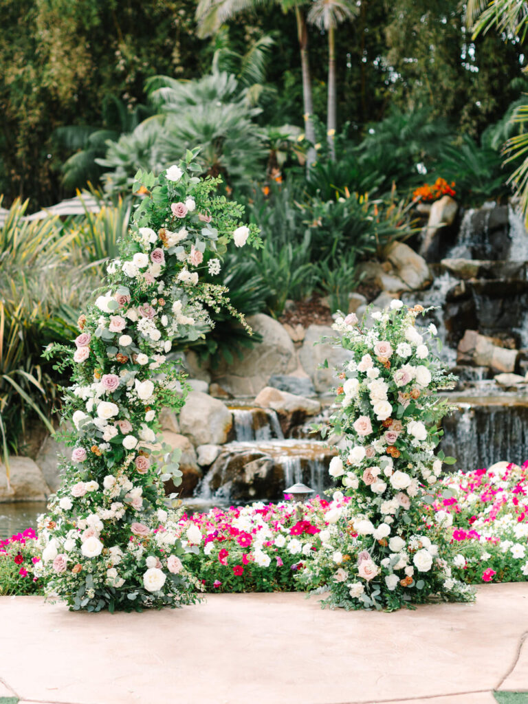 Lush garden scene features two tall floral arrangements with pink and white roses, set against a backdrop of a stone waterfall and vibrant greenery.