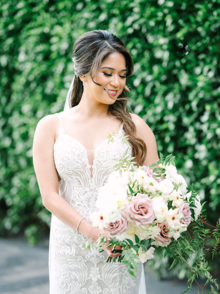 Bride in a lace wedding dress holds a bouquet of pastel flowers and greenery, smiling softly.