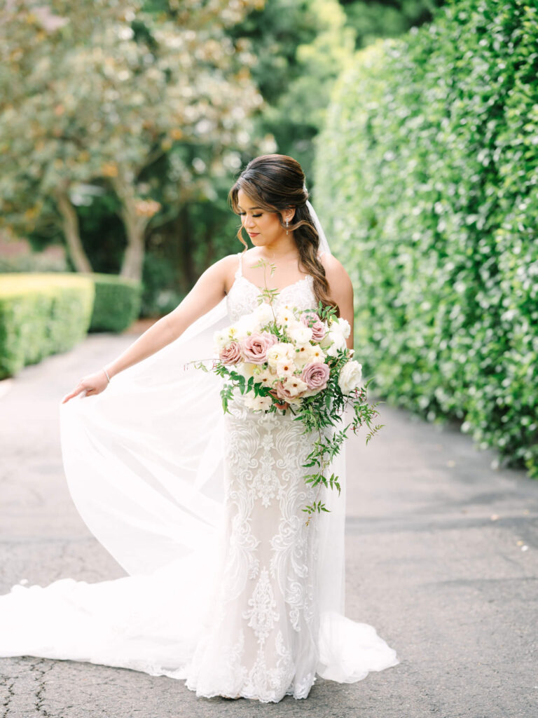 Bride in an elegant lace gown holds a bouquet of pink and white flowers on a garden path, surrounded by lush greenery.