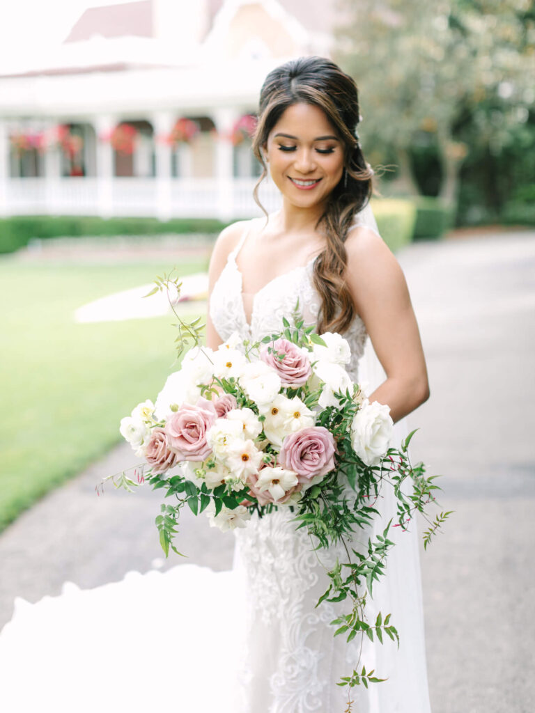Bride in a white lace gown holding a bouquet of pink and white roses, smiling outdoors in a garden setting near a charming house.