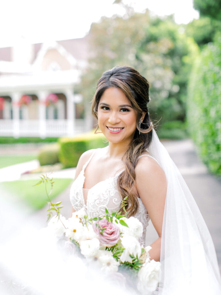 Bride in a white lace gown holding a bouquet of pink and white roses, smiling outdoors in a garden setting near a charming house.
