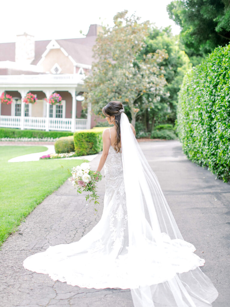 A bride in a flowing white gown and long veil walks on a garden path, holding a bouquet.