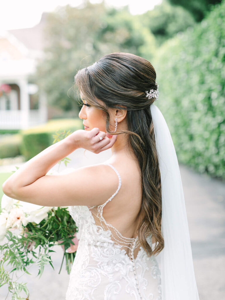 Bride in a lace wedding dress and veil stands outdoors, holding a bouquet. Her hair is elegantly styled with a jeweled clip.