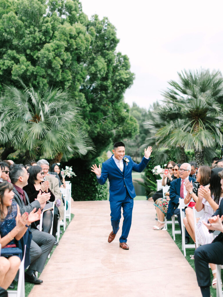 A smiling man in a blue suit enthusiastically walks down an outdoor aisle surrounded by clapping guests.