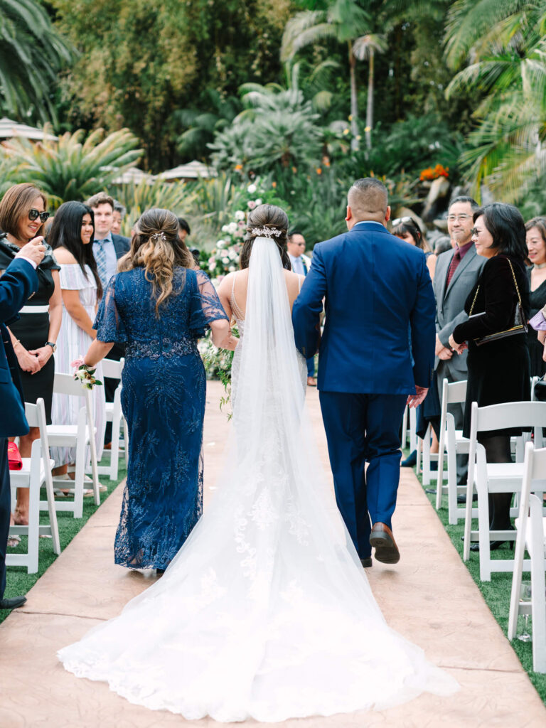Bride in a flowing white gown and veil walks down a lush outdoor aisle, flanked by two people in blue.