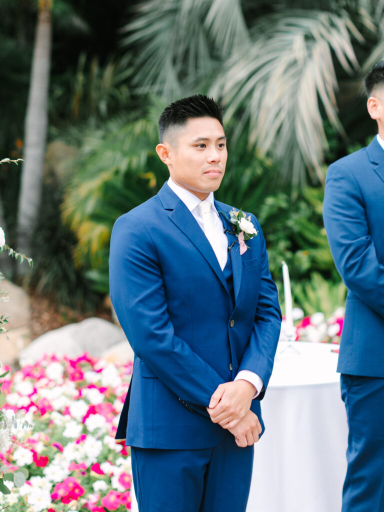 An emotional groom in a blue suit stands outdoors, next to colorful flowers and lush greenery.