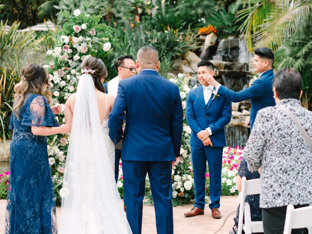A wedding ceremony outdoors with a floral arch. The groom crying upon seeing his bride.