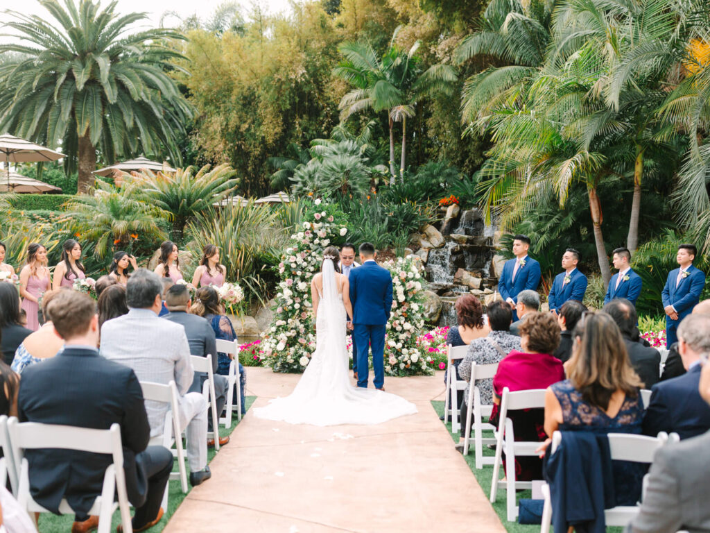 A bride and groom stand at an outdoor altar adorned with flowers, surrounded by a lush garden and waterfall.