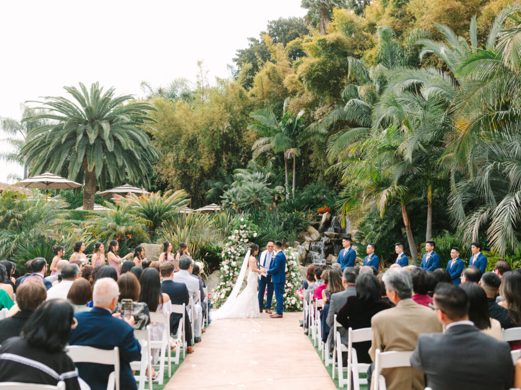 A couple stands at an altar exchanging vows in a lush garden setting.