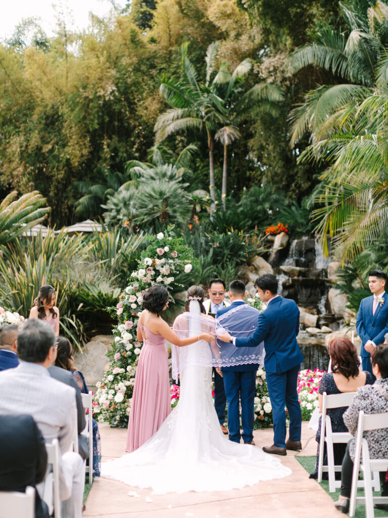 A wedding ceremony in a lush garden setting with groom and bride are wrapped in a veil.