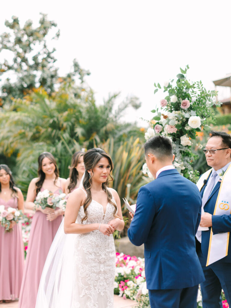 A bride and groom exchange vows outdoors. The bride in a white gown looks at the groom in a blue suit.