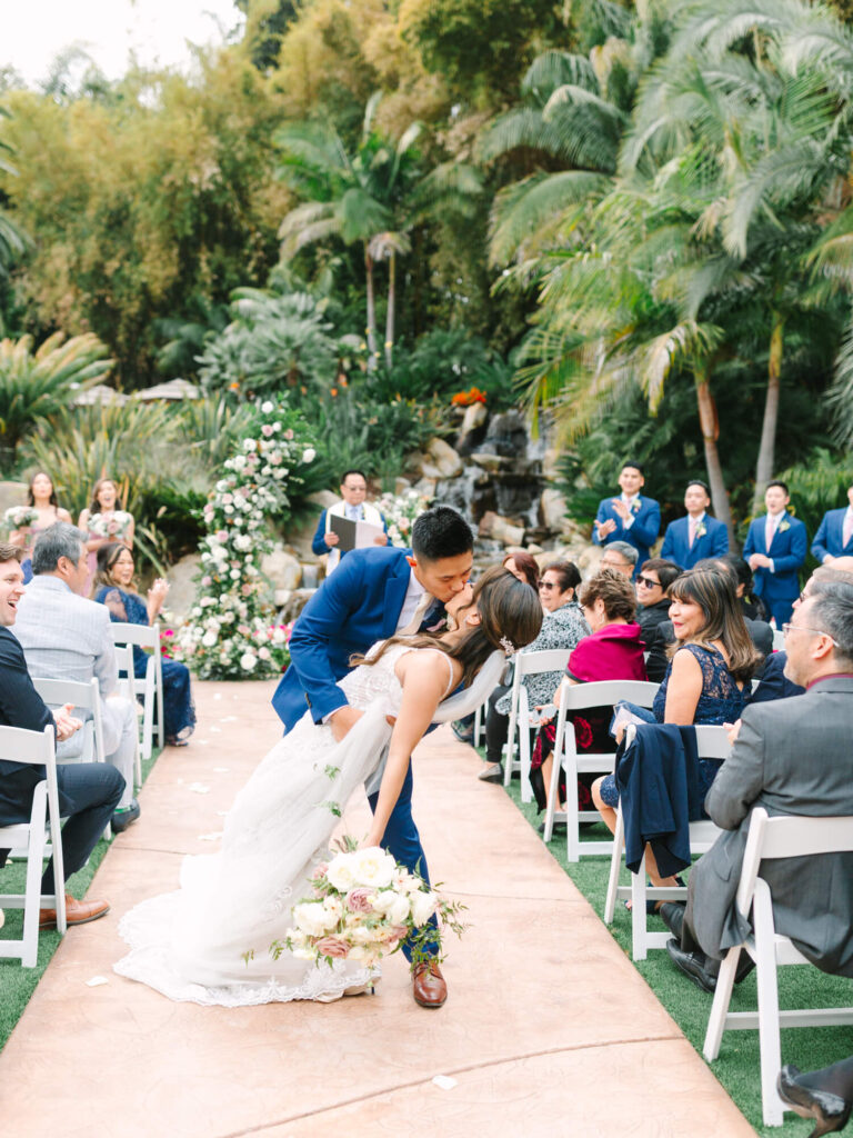 A groom in a blue suit kisses the bride, leaning her back gently as they exit the ceremony.